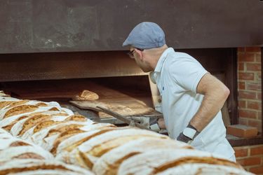 Ein Mann schiebt mit einem Brotschieber ein Brot in einen großen Backofen. Sehr viele fertig gebackene Brote liegen seitlich vor dem Backofen.