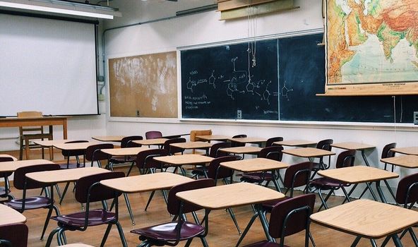 Klassenraum mit vielen SchulTischen und Stühlen. Seitlich Hängt eine große Tafel und ein Teil einer Weltkarte.