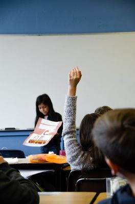 Schüler sitzen in mehreren Tischreihen in einem Klassenraum. Vorne steht ein Mädchen und zeigt ein Plakat. Eine Schülerin meldet sich. 