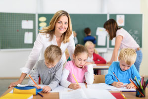 3 Kinder sitzen nebeneinander an einem Tisch und malen. Eine Lehreren schaut ihnen über die Schulter und lächelt in die Kamera. im Hintergrund sind eine große Tafel, weitere Kinder und eine Lehrerin.