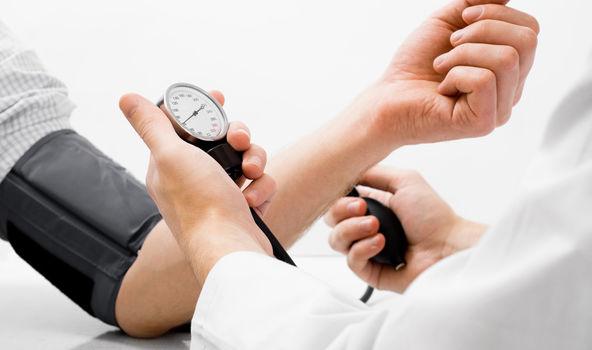 An einem Arm ist ein Blutdruckmessgerät angelegt. Eine weitere Person mit weißem Arztkittel hält in der einen Hand die Pumpe und in der anderen Hand die Anzeige des Messgeräts. 