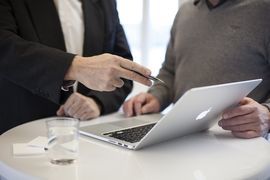 2 Personen, von den man die Gesichert nicht sieht, stehen vor einem Laptop. Der Laptop steht auf einem Stehtisch. Eine Person zeigt der anderen Person durch eine Handbewegung etwas auf dem Bildschirm.