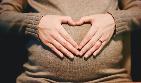 Bauch einer schwangeren Frau. Sie hat ihre Hände auf den Bauch gelegt, sodass die Form eines Herzes entsteht.