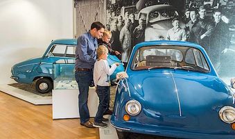 Vater mit 2 Kinder ist in einem Museum. Dort sind 2 Oldtimer ausgestellt. Eines der Autos schauen sie sich näher an.