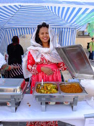 Eine Frau mit bunter Bekleidung und Kopfbedeckung steht an einem Stand und zeigt Essen.