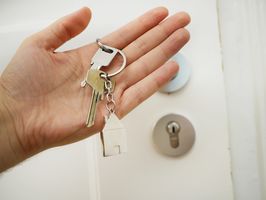 Ein Schlüsselbund mit 2 Schlüsseln und einem Metall-Anhänger in Form eines Hauses hängt am Zeigefinger einer Hand. Im Hintergrund ist das Schloss einer weißen Tür.