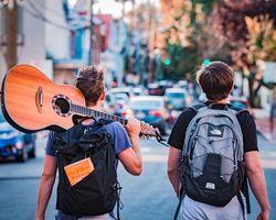 2 Jugendliche mit Rücksack gehen an einer stark befahrenen Straße entlang. Einer der beiden trägt eine Gitarre über der Schulter.