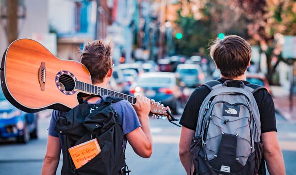 2 Jugendliche mit Rücksack gehen an einer stark befahrenen Straße entlang. Einer der beiden trägt eine Gitarre über der Schulter.