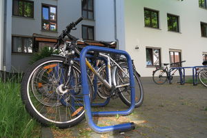 Mehrere Fahrräder an einem Fahrradständer. Im Hintergrund ein großes Haus, vor dem weitere Fahrradständer stehen. Dort parken weitere Fahrräder.