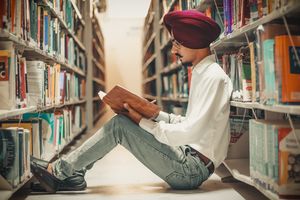 Junger Mann mit Turban sitzt in einer Bibliothek am Boden zwischen zwei großen Regalen mit sehr vielen Büchern. Er macht Notizen in einem Buch.