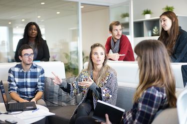 6 junge Erwachsene sitzen und stehen um einem runden Tisch. Sie diskutieren miteinander. Auf dem Tisch steht ein Laptop. Mehrere Zettel und ein Stift liegen daneben. Eine junge Frau hält ein Buch in der Hand.