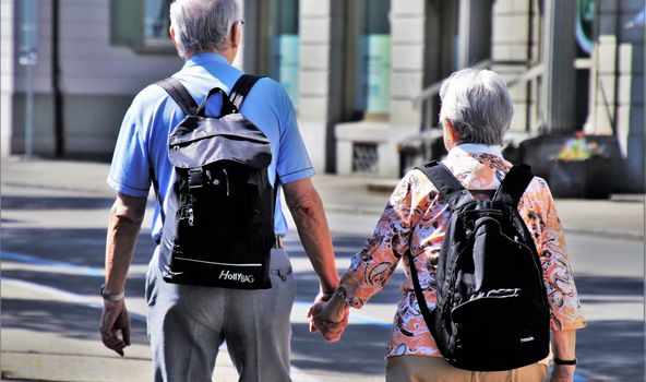 Ein älterer Mann hält die Hand einer älteren Frau. Beide laufen mit Rucksäcken die Straße entlang.