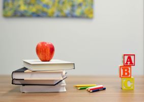 Mehrer Bücher liegen aufeinander auf dem Tisch. Auf den Büchern liegt ein Apfel. Neben den Büchern liegen Buntstifte. Neben den Buntstiften sind 3 Baukötzer mit den Buchstaben A, B und C aufeinander gestapelt. 