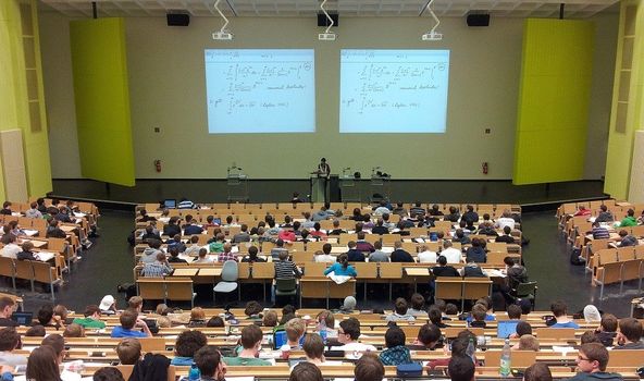 In einem Hörsaal sitzen viele Studenten. Vorne präsentiert ein Professor 2 Folien, die groß an der Wand hinter ihm zu sehen sind.